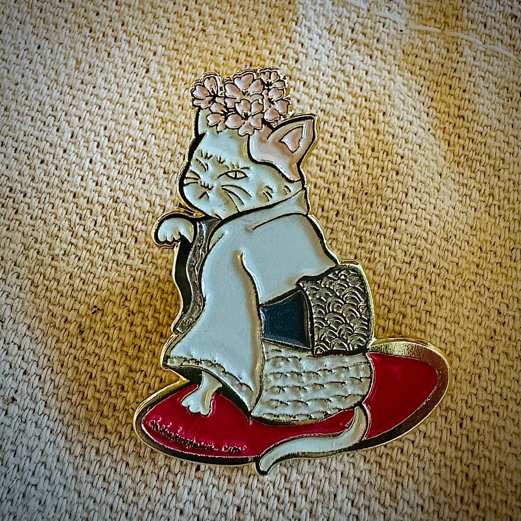 Close-up of a gold enamel pin featuring a cat in a kimono with a cherry blossom crown, standing on a red geta sandal