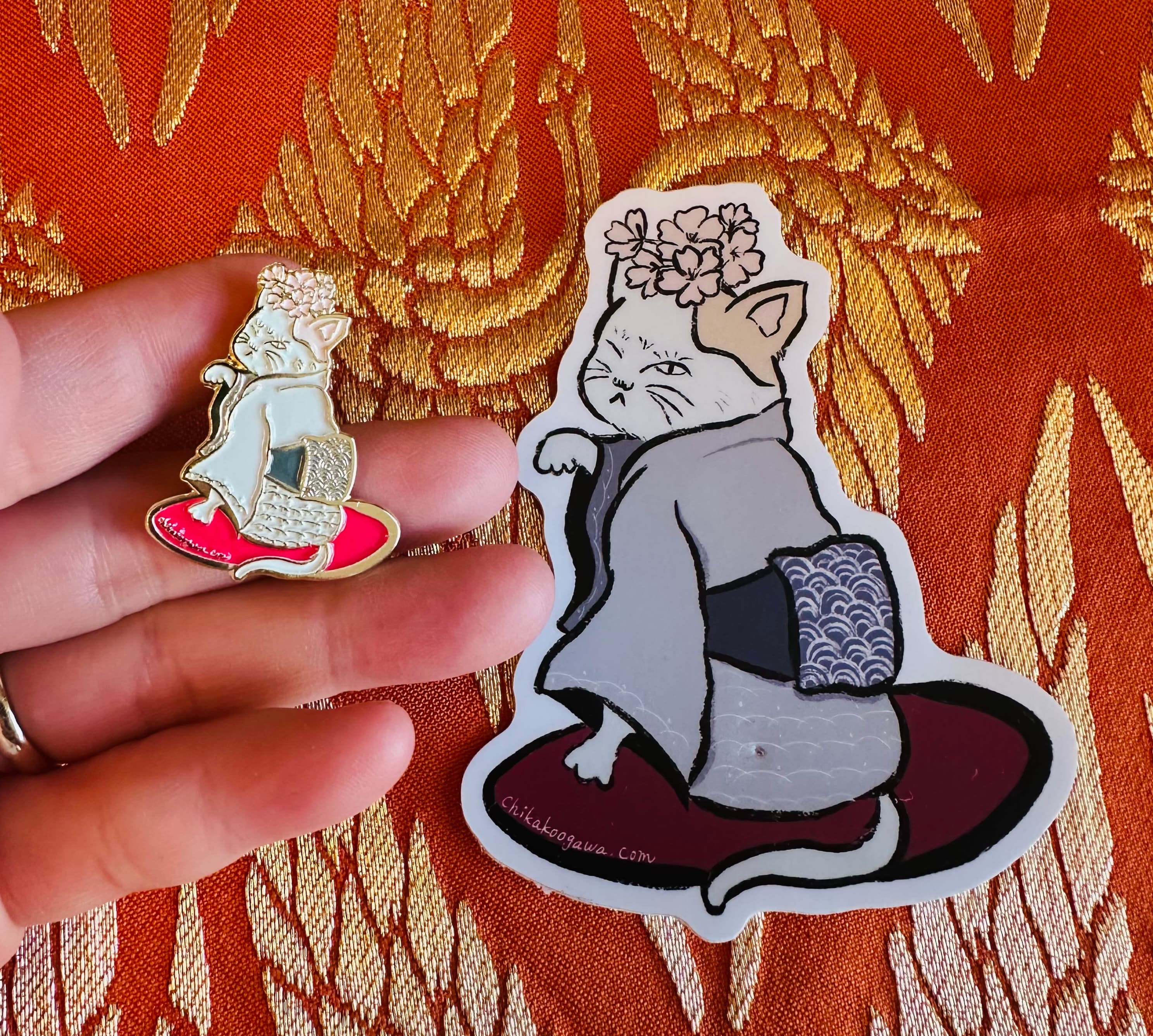 A hand holding the kimono cat enamel pin next to a matching vinyl sticker of the same design, on a gold and orange brocade fabric