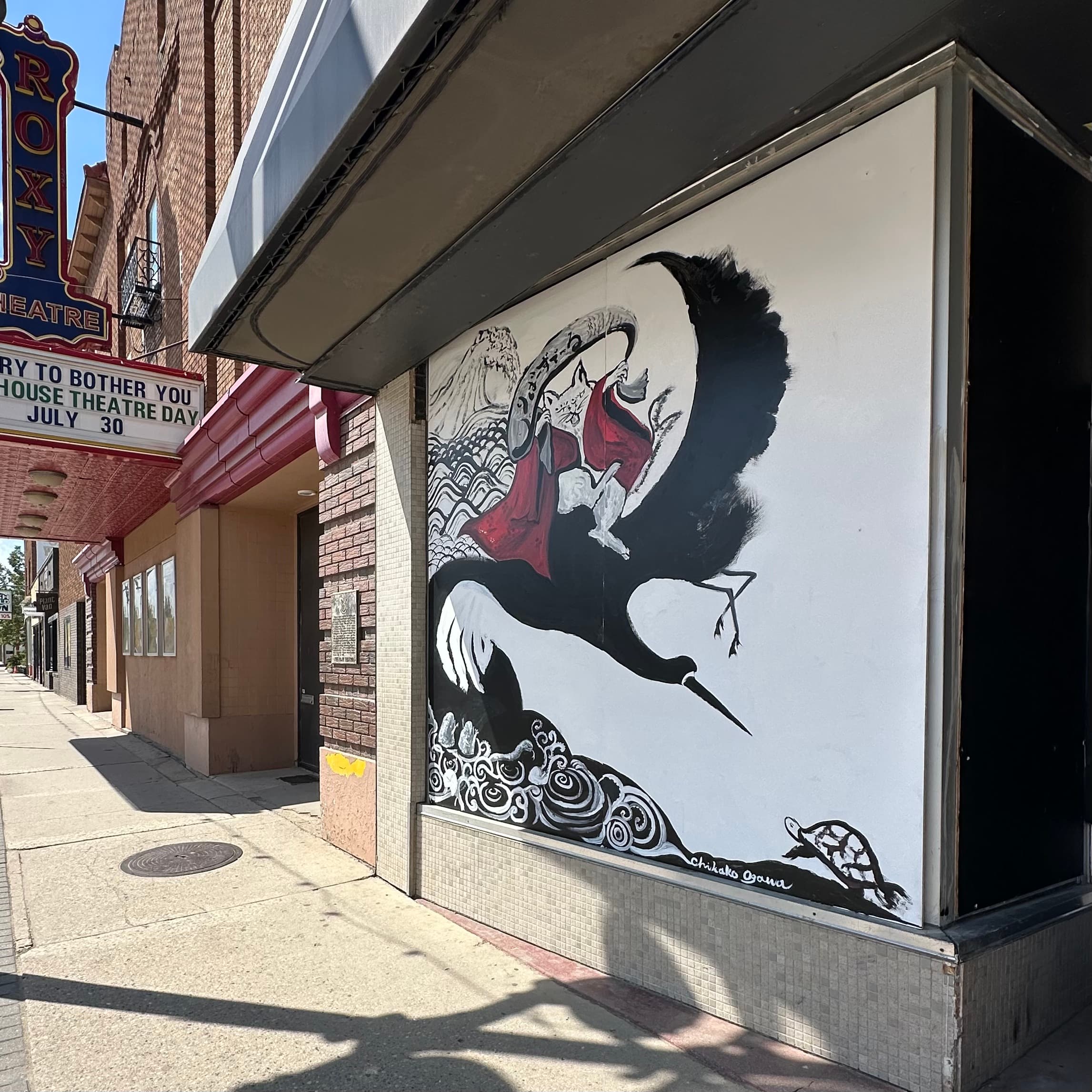 Finished storefront mural near the Roxy Theatre in Saskatoon showing a black crane carrying a cat wrapped in red cloth above waves and a turtle, signed Chikako Ogawa
