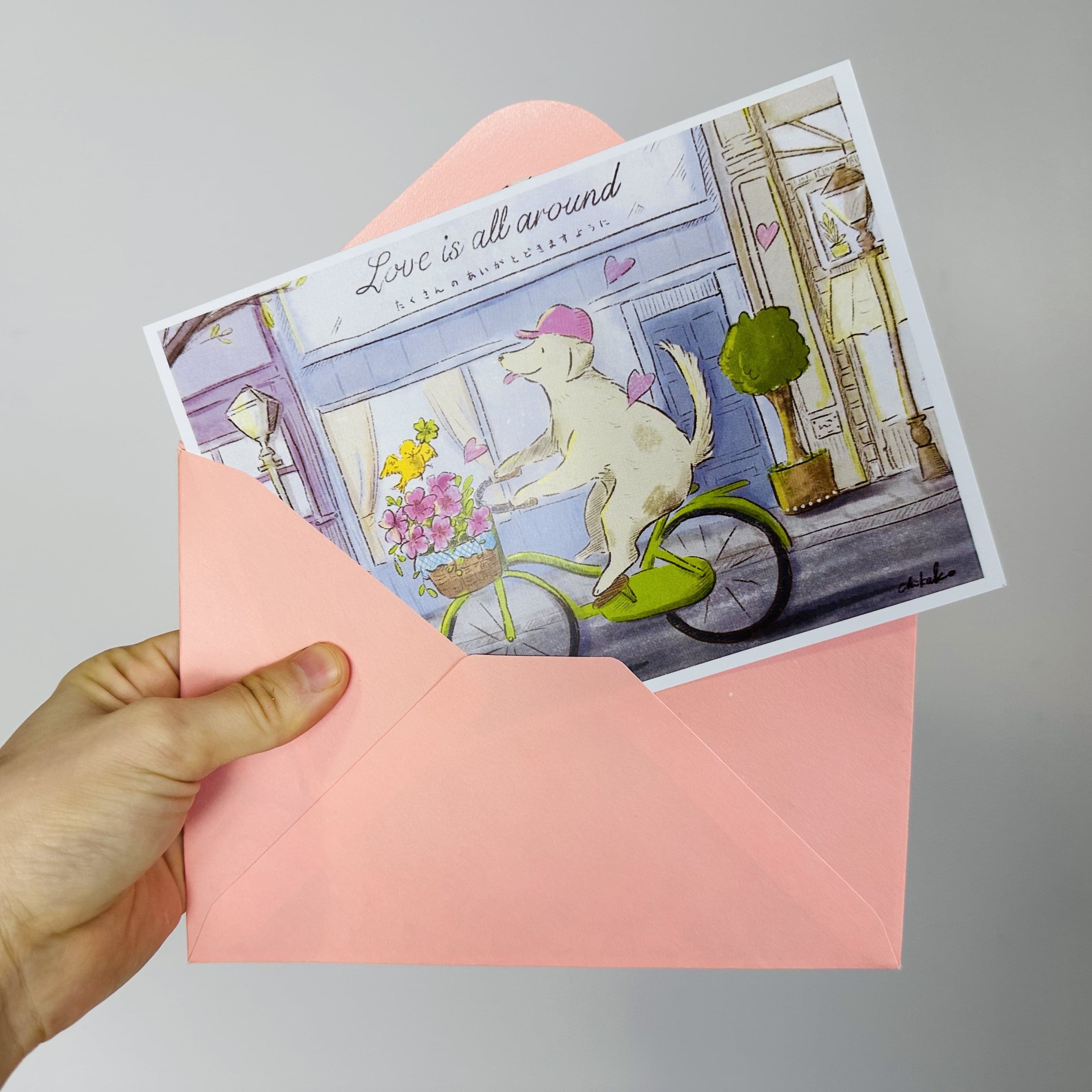 A hand holding an envelope with a postcard in it that says Love is All Around. There is a dog riding a bicycle in front of a city shop.
