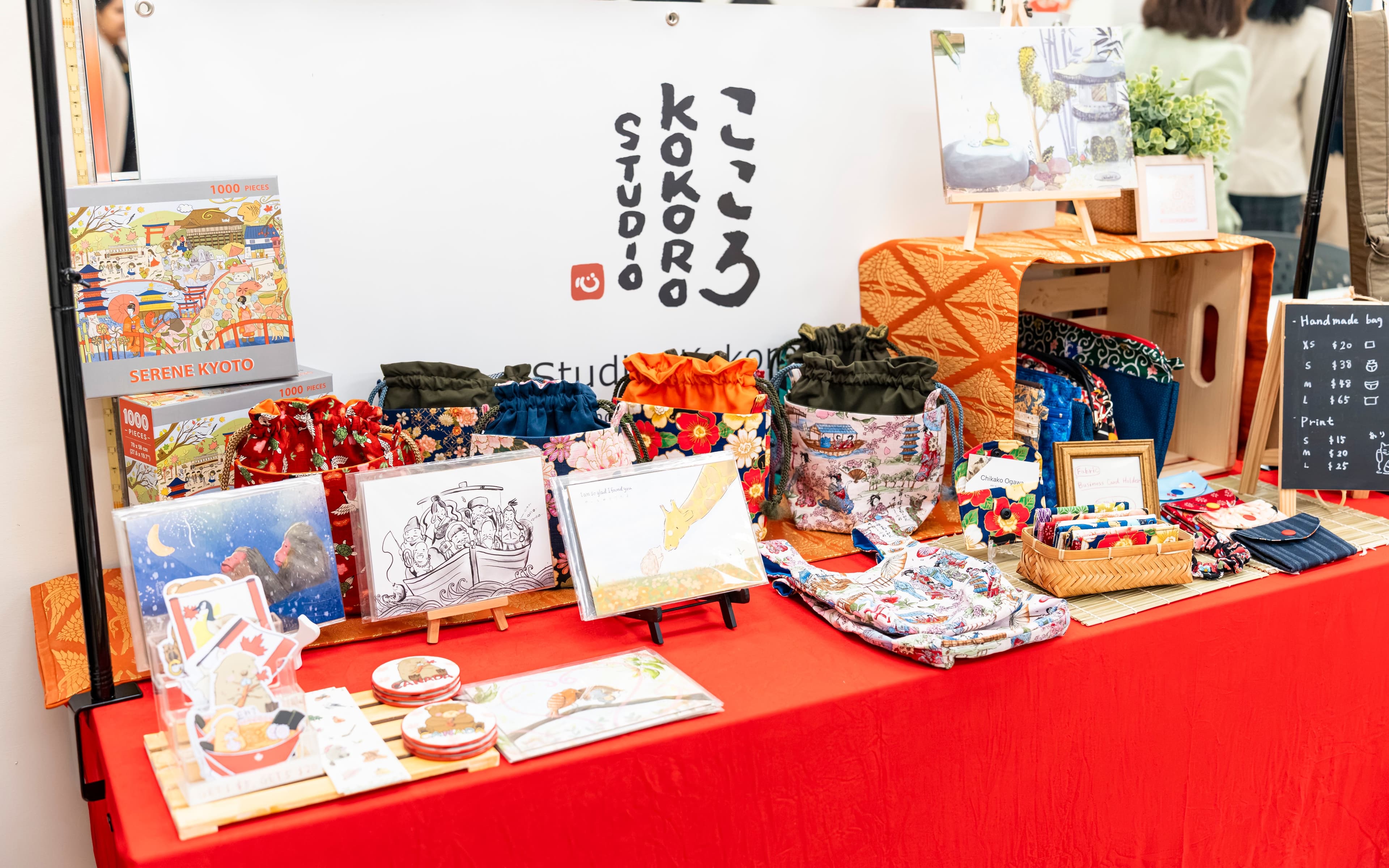 Studio Kokoro market table displaying the logo signage alongside puzzles, drawstring pouches, greeting cards, stickers, and other branded merchandise