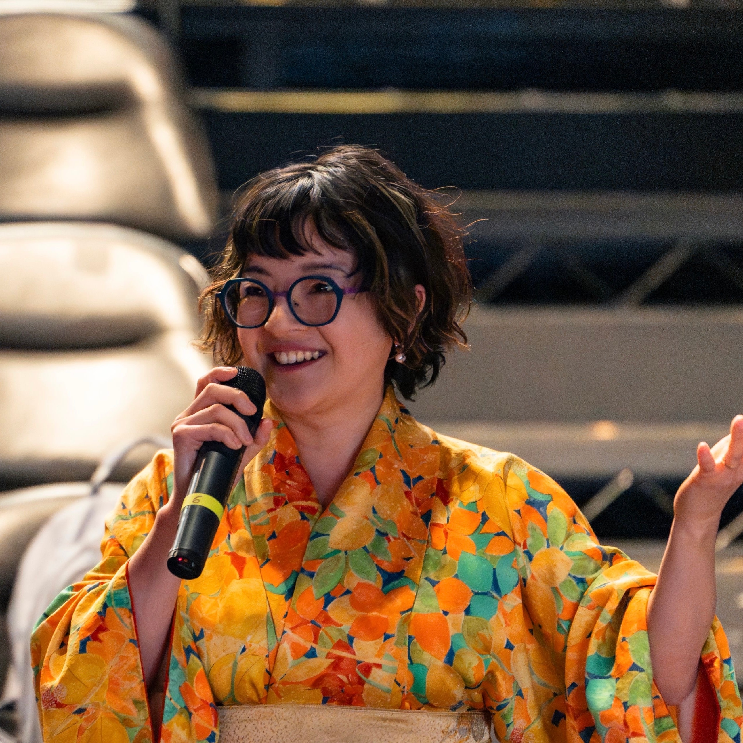 Chikako Ogawa in a floral kimono smiling and speaking into a microphone at an event