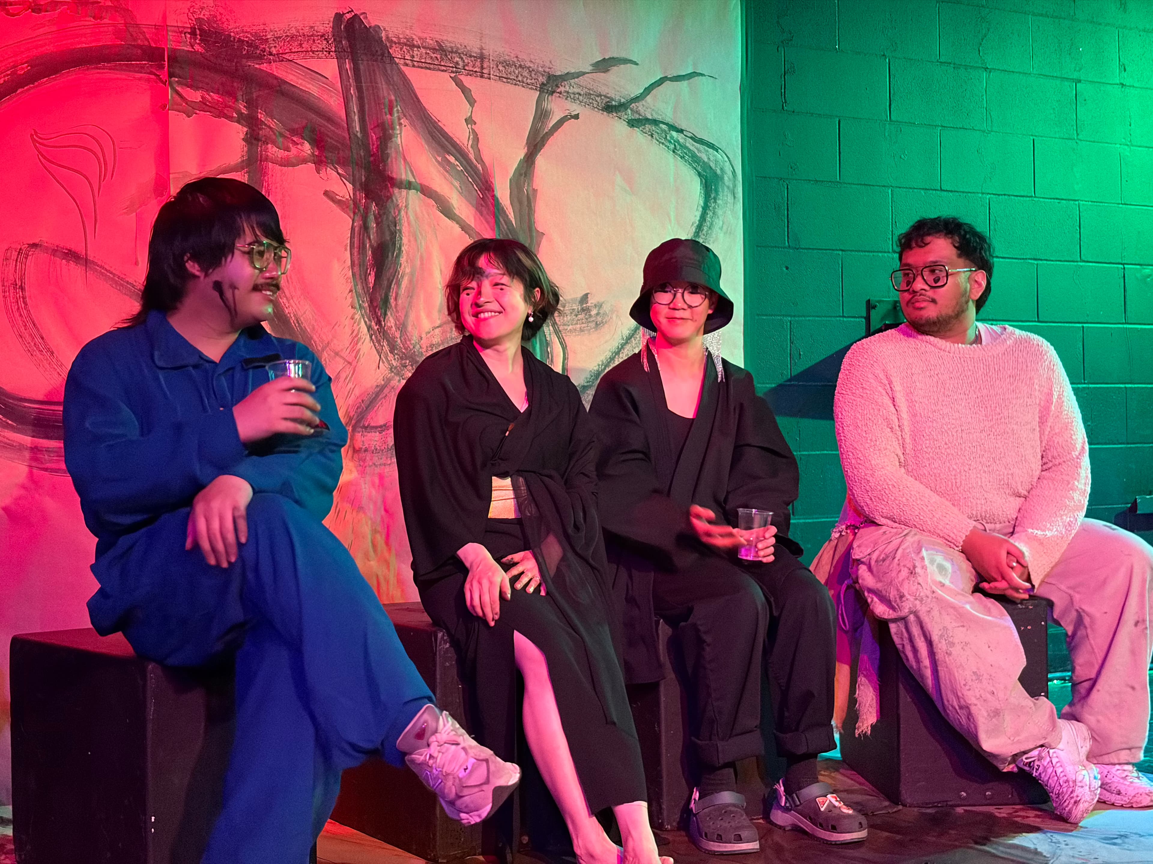 The four Currents collaborators sitting together on stage in front of a large sumi ink mural, lit in red and green stage light