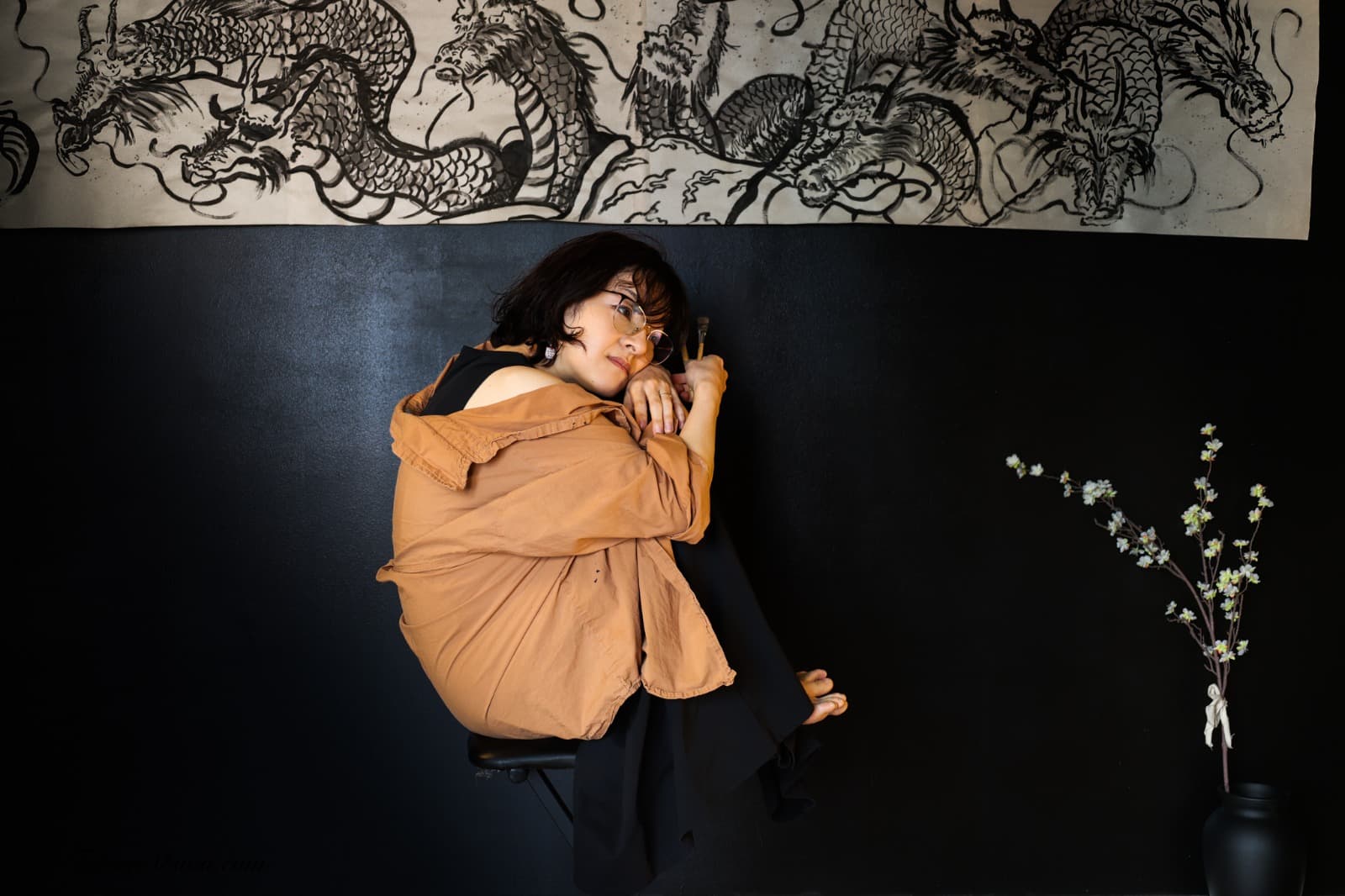 Chikako sitting curled up against a black wall beneath a large ink painting of dragons, with a cherry blossom branch in a vase beside her