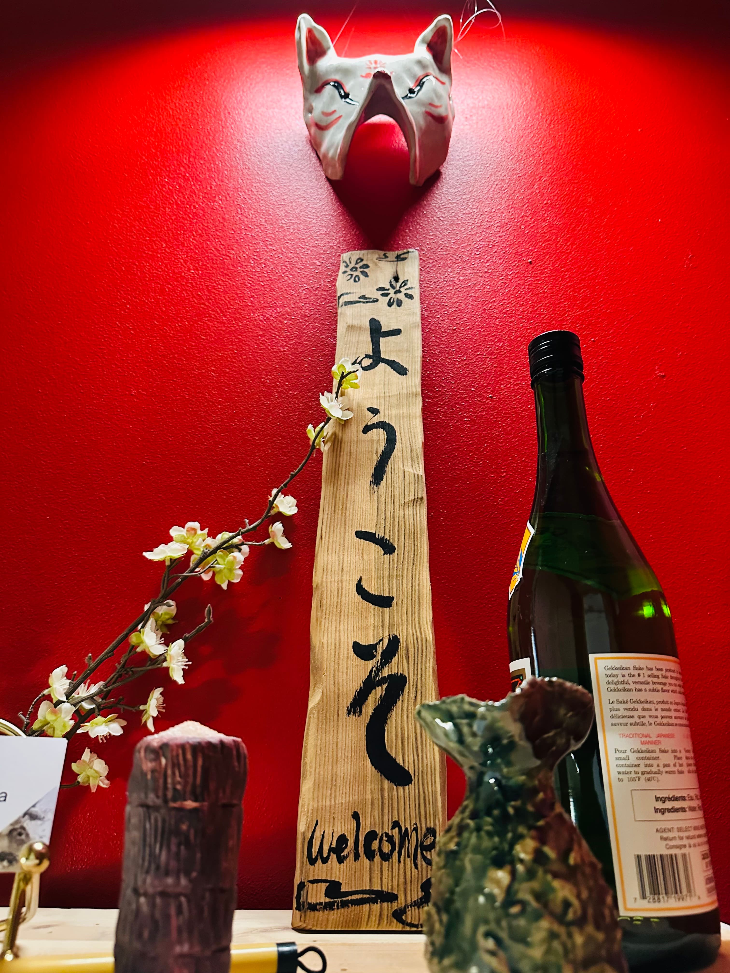 Ceramic kitsune mask mounted on a red wall above a hand-painted wooden sign reading 'ようこそ Welcome', with a sake bottle and cherry blossom sprig nearby