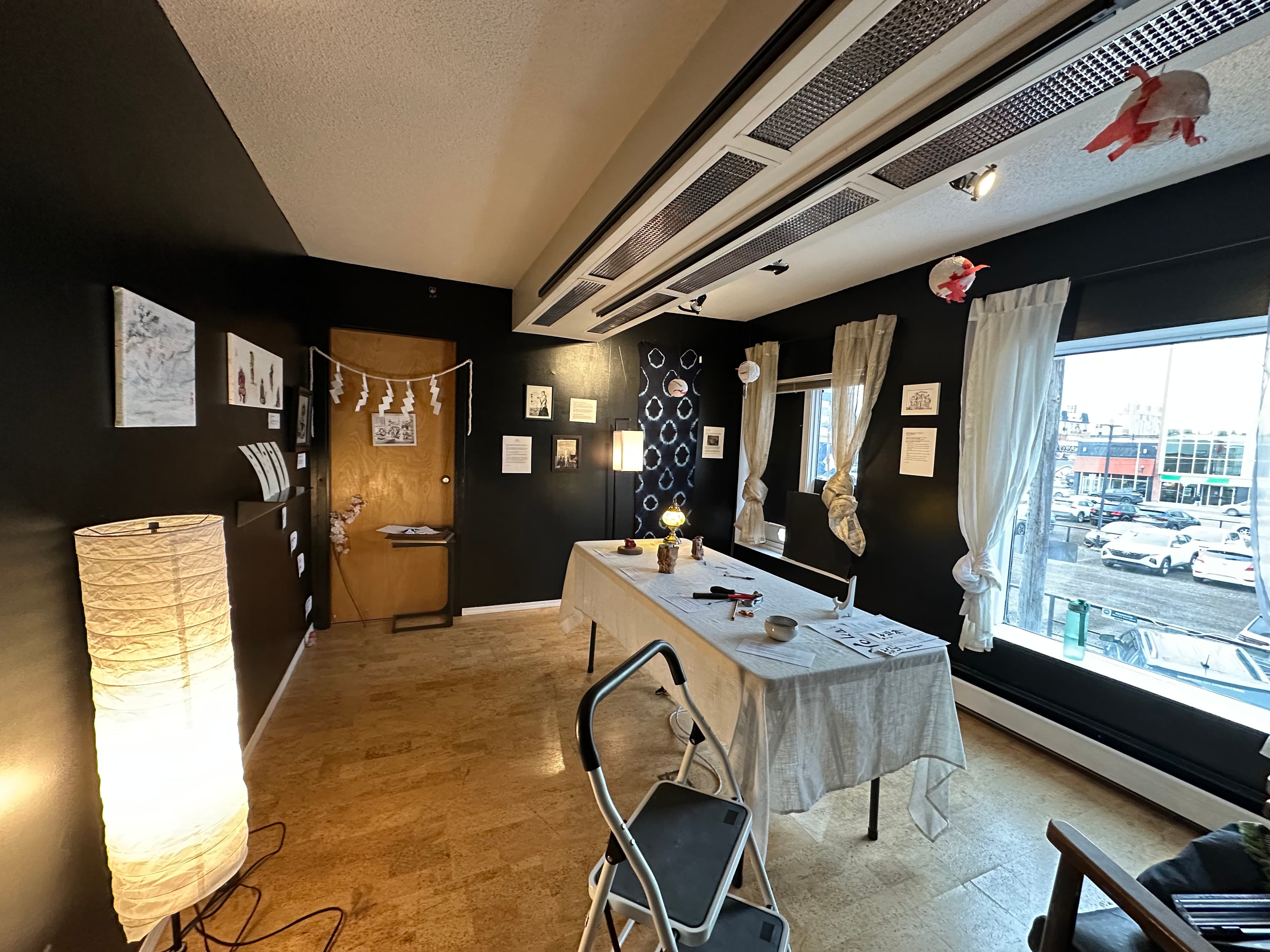 Wide view of the exhibition space with black walls hung with framed artwork, a paper lantern floor lamp, and a linen-covered table