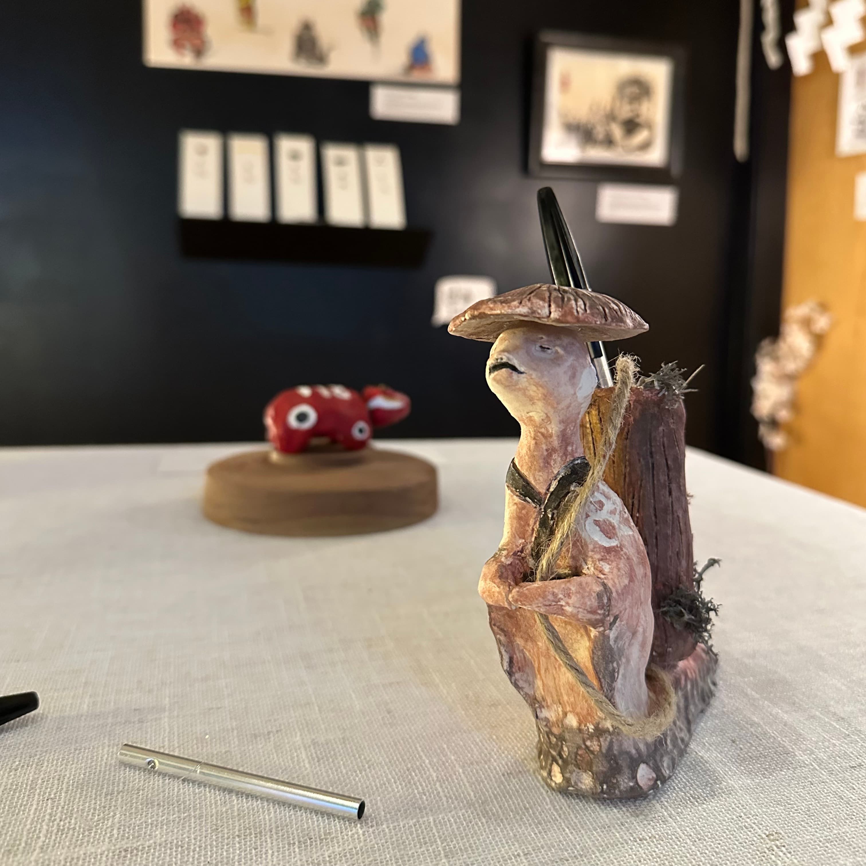 Ceramic yokai figurine wearing a mushroom cap, seated against a tree stump on a table, with a small red creature sculpture in the background