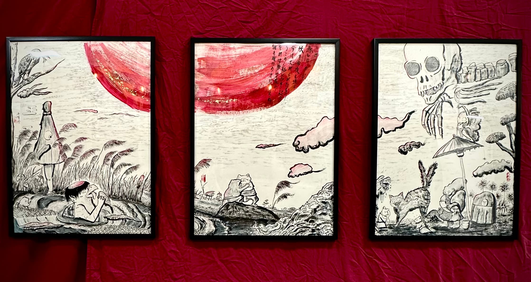 Three framed ink paintings on a red fabric backdrop depicting yokai scenes: a boy bathing near a scarecrow spirit, a toad-like yokai on a rock, and a skeletal hand looming over cats and an umbrella figure