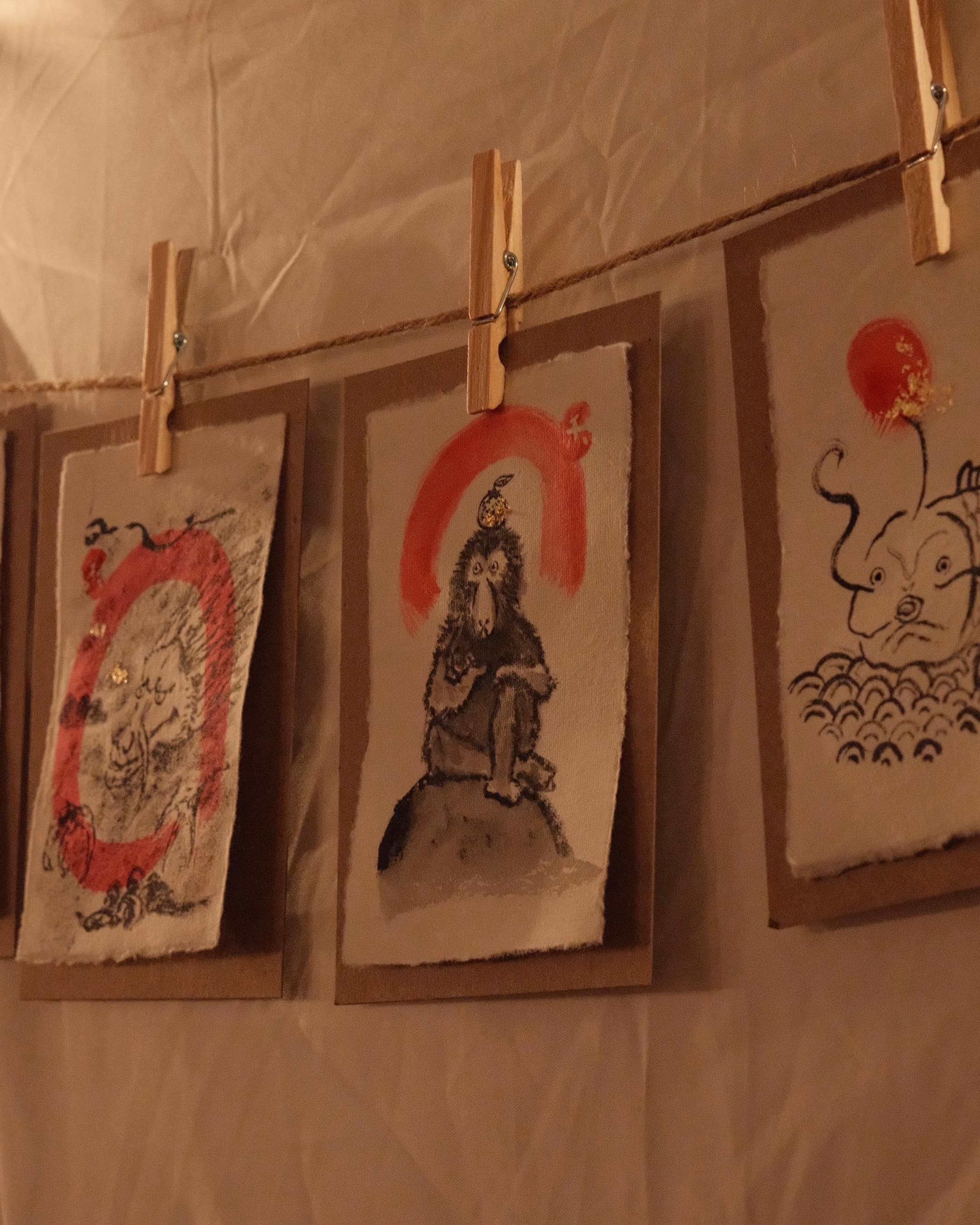 Small ink and watercolor yokai sketches clipped to a hanging line, including a coiled red dragon, a monkey under a red arc, and a horned creature