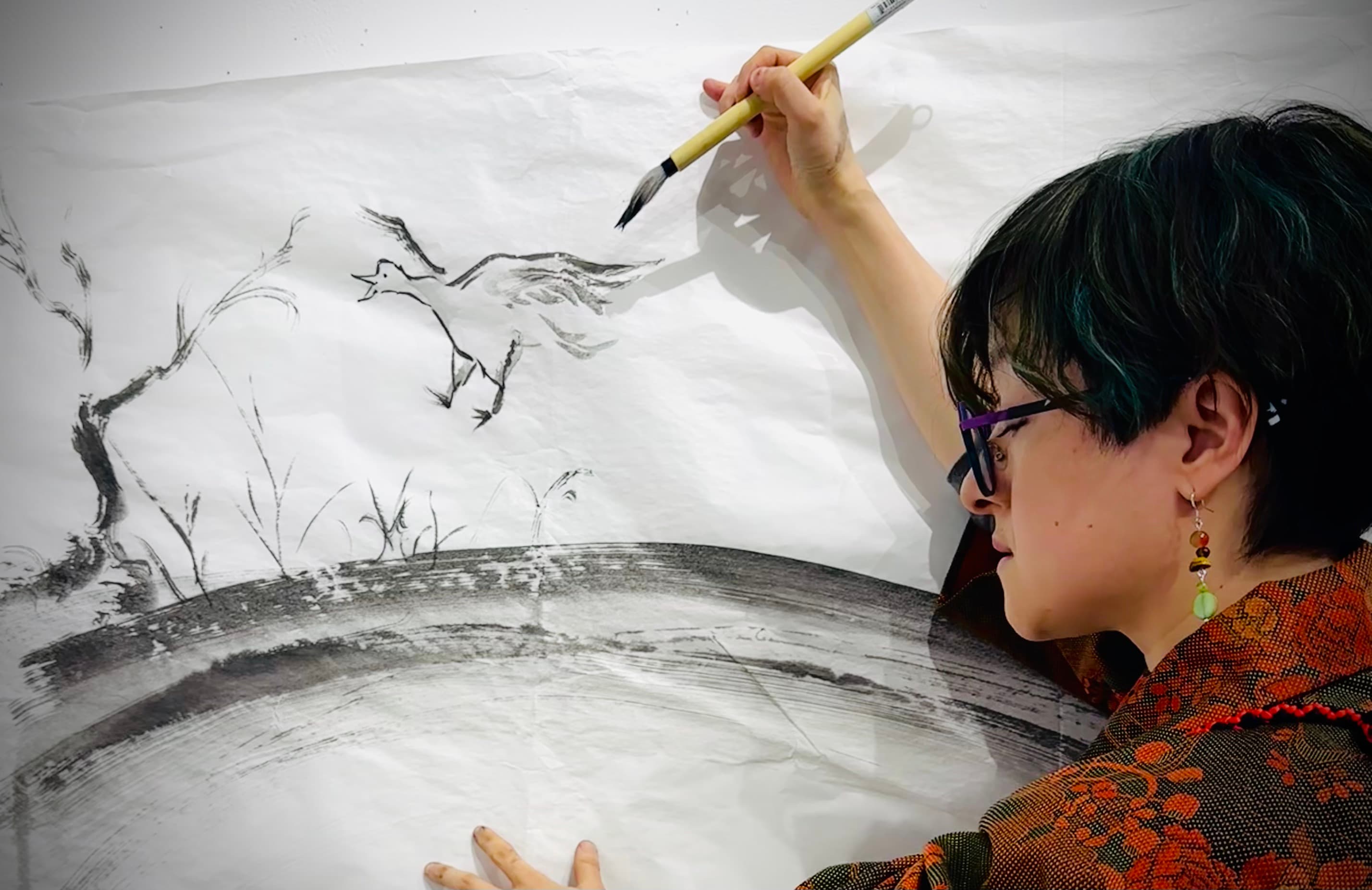 Chikako painting a large sumi ink piece of a bird and reeds on a wall-mounted sheet of paper