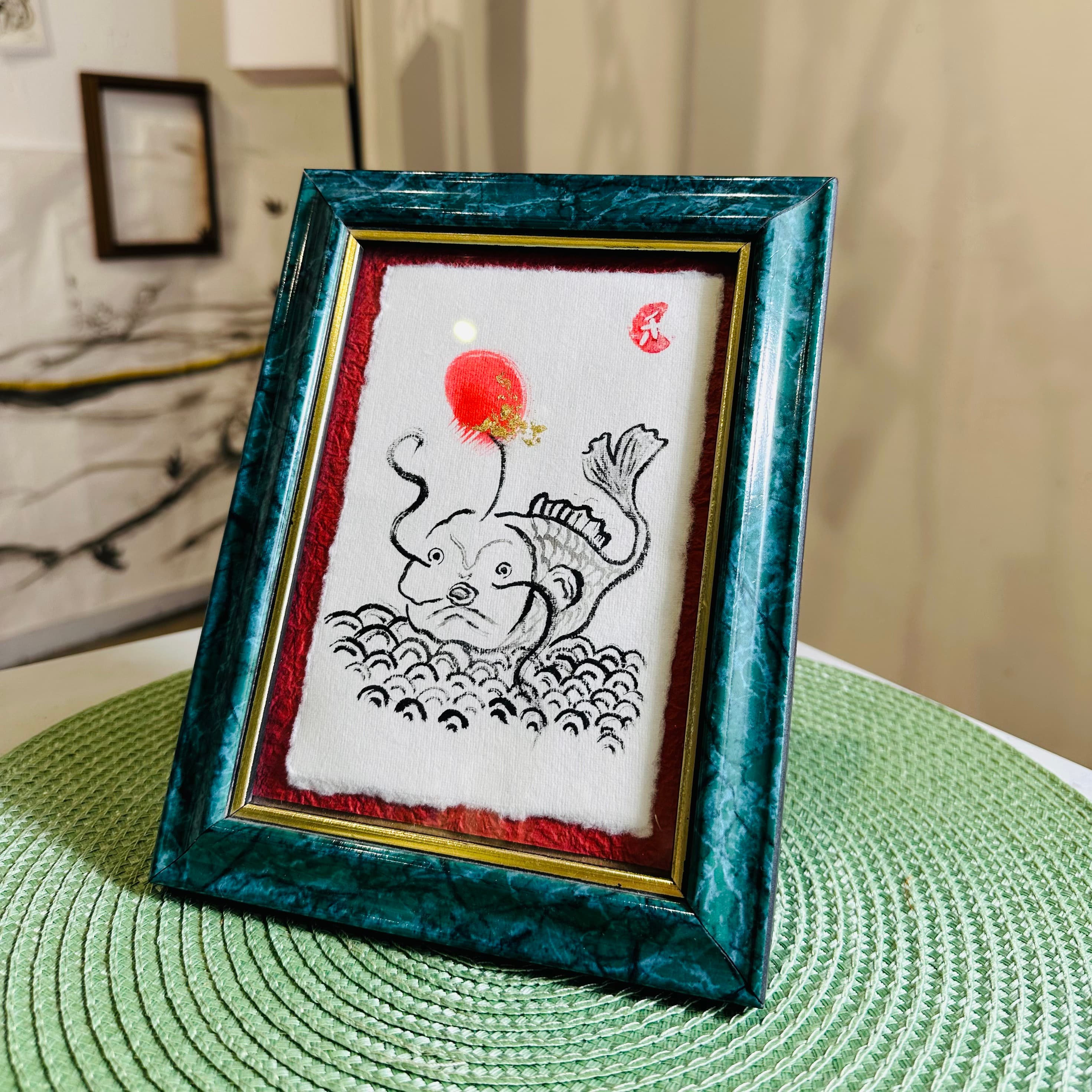 Framed ink painting of a fish leaping from waves beneath a red sun, in a dark green marbled frame