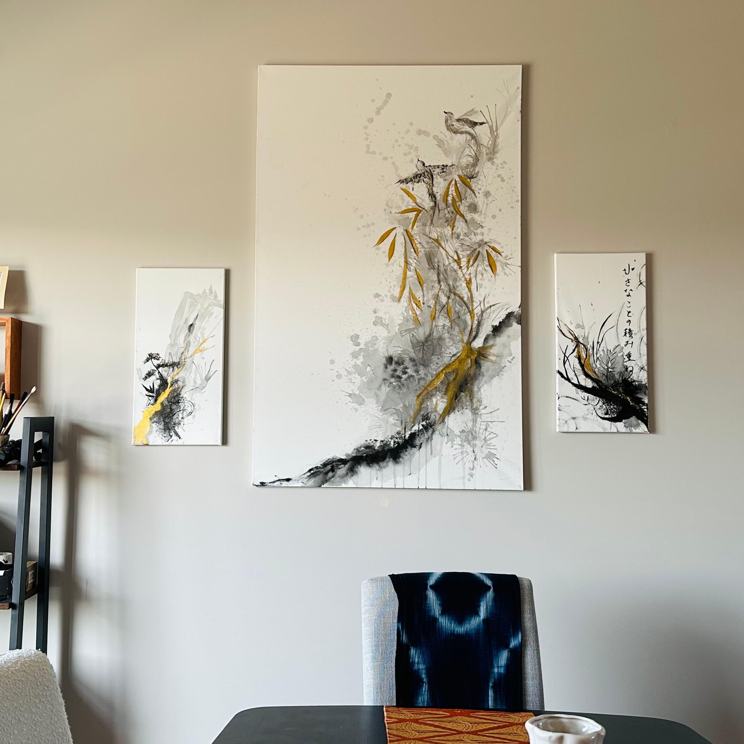 Three ink paintings on a dining room wall: a large piece of birds perched on gold bamboo branches, flanked by two smaller abstract ink and gold works