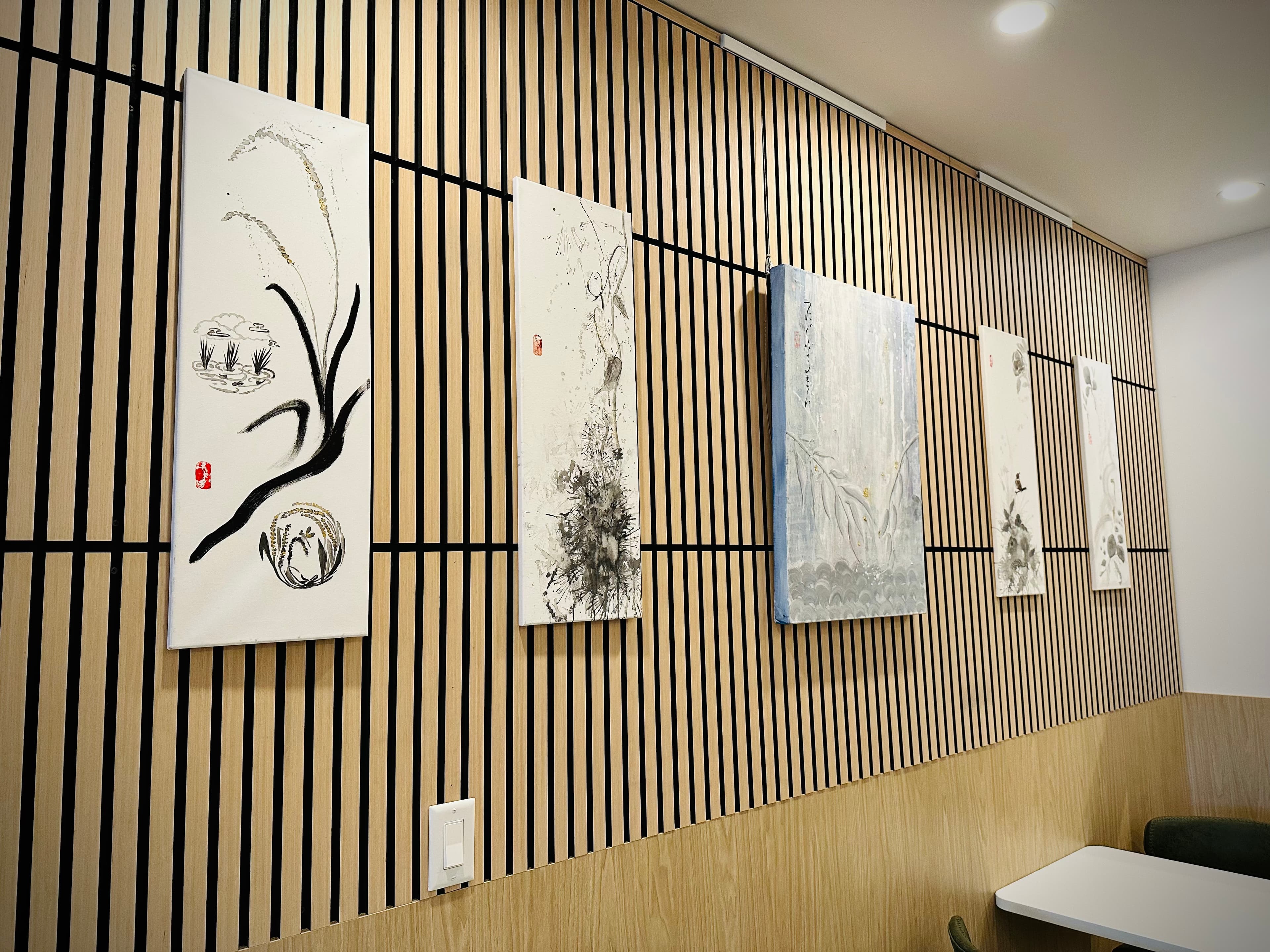 Row of ink paintings hung on a wood-slat gallery wall, including a branch and reed piece, an ink splatter abstract, and a pale blue washed work